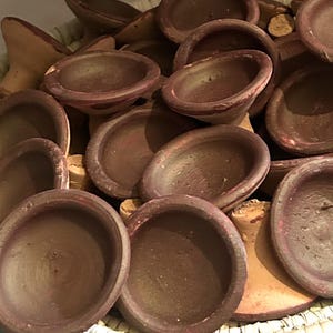 Puede incluir: Una cesta tejida llena de pequeños cuencos de terracota marrones. Los cuencos tienen una forma poco profunda y redondeada y están apilados. La cesta tiene un patrón tejido natural con un borde rosa y morado.
