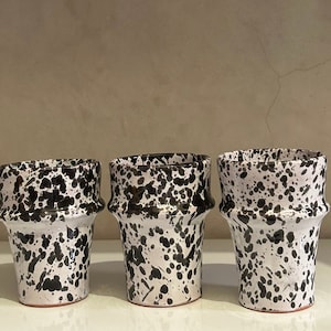 Peut inclure: Trois vases en céramique avec une base blanche et un motif éclaboussé noir. Les vases ont une forme unique avec un dessus évasé et une surface texturée. Les vases sont disposés en rangée sur une surface blanche.