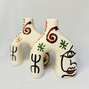 Peut inclure: Deux vases en céramique blanc cassé de forme arquée. Chaque vase est peint à la main avec des motifs abstraits noirs, rouges et verts, dont un visage stylisé. Les vases ont une finition brillante et sont posés sur un fond uni.