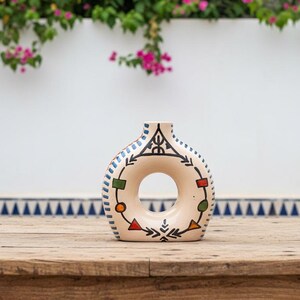 Peut inclure: Un vase en céramique beige avec un centre circulaire et un col étroit. Le vase est décoré de motifs géométriques noirs, bleus, rouges, verts et oranges. Le vase est exposé sur une table en bois.