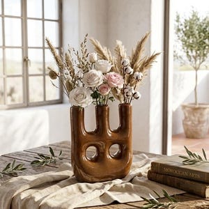 Peut inclure: Un vase en céramique marron à trois ouvertures, rempli de roses blanches et roses, de coton et d'herbes séchées. Le vase est posé sur une table en bois rustique avec un chemin de table en lin. Deux livres et des brins de verdure complètent le décor.