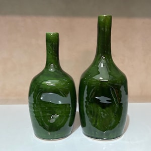 Pode incluir: Duas garrafas de cerâmica verdes com acabamento brilhante. As garrafas têm uma forma clássica com gargalo estreito e corpo arredondado. As garrafas são de tamanhos diferentes. Elas são exibidas em uma superfície branca. Provavelmente são decorativas.