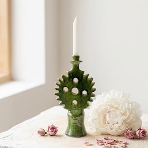 Peut inclure: Un bougeoir en céramique vert avec une bougie blanche, présentant un design décoratif unique avec des découpes circulaires. Une fleur blanche et des boutons de rose séchés sont disposés à proximité, créant une présentation naturelle et élégante.