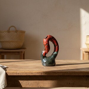 Peut inclure: Un vase en céramique abstrait avec un motif tacheté rouge et noir. Le vase a une forme incurvée et organique et est posé sur une table en bois. Des paniers en osier sont en arrière-plan.