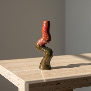 Peut inclure: Un vase en céramique artisanal avec un design unique et sinueux. La partie supérieure est rouge vif avec une texture tachetée, passant à une base vert foncé. Le vase est présenté sur une table de couleur claire.