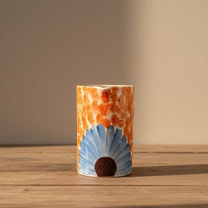 Peut inclure: Un vase cylindrique en céramique avec un motif peint à la main. Le vase présente un motif tacheté orange et un motif floral bleu avec un centre marron. Le vase est posé sur une surface en bois.