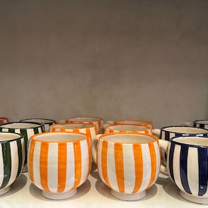 Peut inclure: Une collection de tasses en céramique rayées de différentes couleurs, dont l'orange, le vert et le bleu. Les tasses ont un fond blanc avec des rayures colorées.