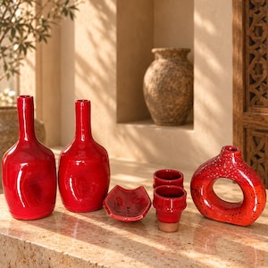 Peut inclure: Une collection d'objets en céramique rouge comprenant deux grandes bouteilles à col étroit, un petit plat carré, deux tasses cylindriques empilées et un vase circulaire en forme de beignet avec une finition texturée noire et rouge.
