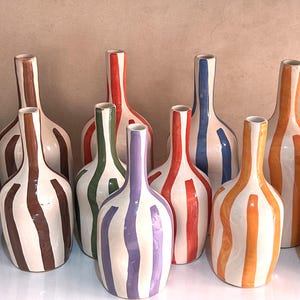 Peut inclure: Ensemble de bouteilles en céramique à rayures verticales de différentes couleurs : violet, marron, vert, rouge, bleu et orange. Les bouteilles ont une base blanche et un col fin, créant un affichage décoratif.