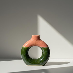 Peut inclure: Un vase en céramique de forme originale en donut. La moitié supérieure est de couleur corail moucheté, tandis que la moitié inférieure est vert foncé. Le vase a une ouverture circulaire au centre et un petit col.