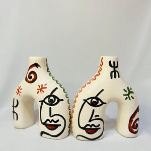 Peut inclure: Deux vases en céramique blanc cassé avec des bases arquées, chacun représentant un visage peint avec des contours noirs, des lèvres rouges et un seul œil. Un vase a des accents verts et rouges, l'autre a des accents rouges et verts. Décoratif et artistique.
