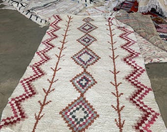 May include: A rectangular off-white rug featuring a central column of diamond shapes in shades of red, blue, and gray. The rug is bordered by two vertical zigzag lines in red and pink, with fringe detailing along the bottom edge of the textile.