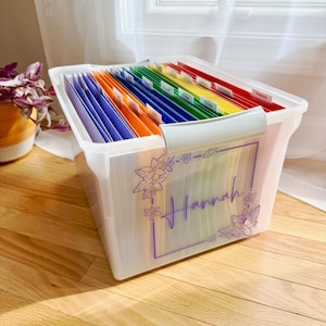 May include: A clear plastic file box filled with colorful file folders. The box has a white handle and a purple floral design with the name "Hannah" on the front. The file folders are labeled with various colors.