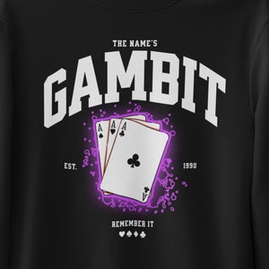 Pode incluir: Camisola de moletom preta com um gráfico branco do texto "The Name's Gambit" com um design de carta de jogo. O texto "Est. 1990" está abaixo do gráfico e o texto "Remember It" está abaixo disso.