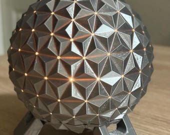 Könnte beinhalten: Eine graue, geometrische Kugellampe mit strukturierter Oberfläche, die auf einem passenden Ständer ruht. Die Lampe strahlt ein warmes, orangefarbenes Leuchten durch die dreieckigen Aussparungen aus. Das Design erinnert an eine geodätische Kuppel.