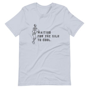 May include: A light blue t-shirt featuring a skeleton graphic and the text "WAITING FOR THE KILN TO COOL."