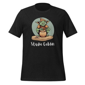 May include: A black t-shirt with a green goblin illustration. The goblin is wearing a brown hat and is working on a pottery wheel. The text "Studio Goblin" is printed below the illustration.