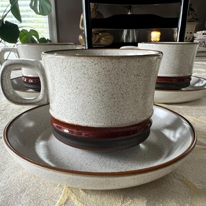 May include: A set of ceramic cups and saucers featuring a speckled off-white finish on the upper portion and a solid dark brown band around the base. Each cup has a wide, squared handle and sits on a matching saucer with a brown rim.