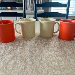 May include: Four ceramic mugs, two in a bright orange and two in a cream colour. The mugs have a classic design with a rounded body and a sturdy handle, ideal for tea or coffee.