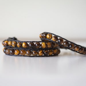 Puede incluir: Dos pulseras de cuero con cuentas de ojo de tigre. Las pulseras son de color marrón y dorado y están superpuestas.