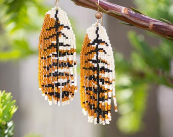 May include: A pair of beaded earrings with a feather design. The earrings are made with white, black, and orange beads. The earrings are hanging from a branch.