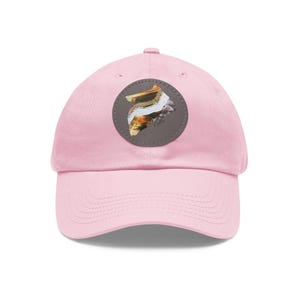 Könnte beinhalten: Eine hellrosa Baseballkappe mit einem kreisförmigen dunkelgrauen Aufnäher auf der Vorderseite. Der Aufnäher zeigt eine abstrakte Grafik in Gold-, Weiß- und Brauntönen, wobei das Wort Collie in kleinen, dunklen Kleinbuchstaben darunter steht.