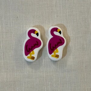 May include: Two flamingo-shaped decorative objects. Each flamingo has a white body, a magenta-colored body and head, a yellow beak and feet, and a black eye. The objects are likely earrings or decorative pins.