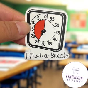 May include: A white sticker with a black border featuring a timer design. The timer shows a red section and the text "I Need a Break". The sticker is held in a hand.