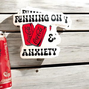 May include: White sticker with black text that reads "RUNNING ON Y & ANXIETY". Two red Coca-Cola cans are pictured in the middle of the sticker.