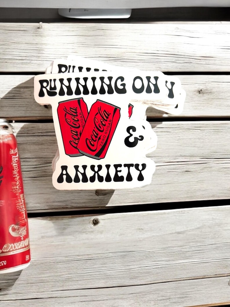 May include: White sticker with black text that reads "RUNNING ON Y & ANXIETY". Two red Coca-Cola cans are pictured in the middle of the sticker.