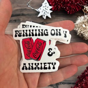 May include: White sticker with black text that reads "Running on Y & Anxiety" with a retro font. Two red Coca-Cola cans are featured on the sticker, one with "Zero Sugar" printed on it.