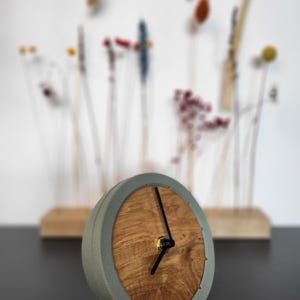 Puede incluir: Un reloj gris y de madera con agujas negras. El reloj está sobre una base de madera.
