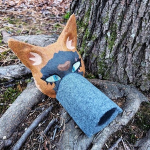 May include: A brown and black felt cat mask with blue eyes. The mask is lying on the ground next to a tree trunk.