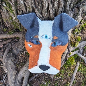 May include: A handmade felt dog mask with a white face, brown and black fur, and blue eyes. The mask has a third eye in the center of the forehead.