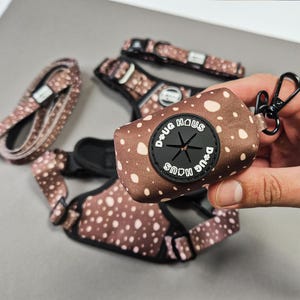 Puede incluir: Un arn&eacute;s y una correa para perro marrones y blancos con un portador de bolsas para caca a juego. El portador de bolsas para caca tiene un c&iacute;rculo negro con el texto "D+UG HAUS" escrito en blanco.