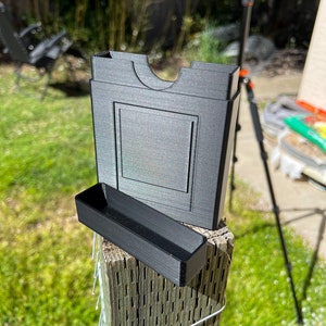 May include: A black 3D printed card holder with a separate tray. The holder has a slot for cards and a raised lip around the top. The tray is rectangular and has a lip on one side. The holder and tray are sitting on a wooden post.