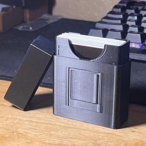 May include: A black, rectangular card holder with a hinged lid, open to the side. The holder has a rectangular design with a recessed square on the front. The holder is on a wooden surface.