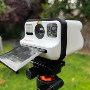 May include: A white Polaroid Now+ instant camera with a black case and a black film pack attached to the side. The camera is mounted on a black tripod with an orange base.