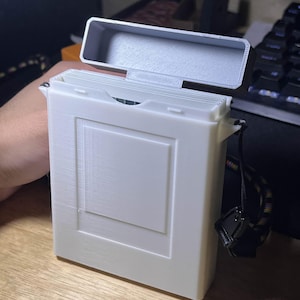 以下が含まれることがあります： 前面に隆起した長方形のデザインが施された、白い長方形のヒンジ付きボックス。蓋は開いており、灰色の内部が見えます。黒とマルチカラーのストラップが取り付けられています。