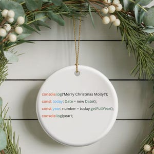 May include: A white ceramic Christmas ornament with gold string, featuring text in a coding style that reads "Merry Christmas Molly!". The ornament hangs against a backdrop of a festive green and white wreath.