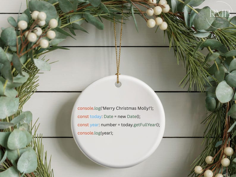 May include: A white ceramic Christmas ornament with gold string, featuring text in a coding style that reads "Merry Christmas Molly!". The ornament hangs against a backdrop of a festive green and white wreath.