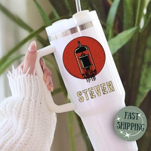 May include: White travel mug with a handle, featuring a red circle with a vintage vacuum tube graphic. The name "STEVEN" is printed below the graphic. The mug has a silver rim and a white lid.