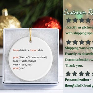 May include: White ceramic Christmas ornament with gold string, displayed in a gift box. The ornament features a Python code snippet with the text "Merry Christmas Mina!" and the current year. Festive holiday decor.