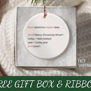 May include: A white ceramic Christmas ornament with a Python code snippet printed on it. The code includes a greeting and the current year. The ornament is in a gift box with a ribbon.