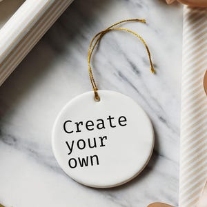May include: A white, round ceramic ornament with the text "Create your own" in black. The ornament hangs from a gold string. The background includes wrapping paper, ribbon, and a marble surface.