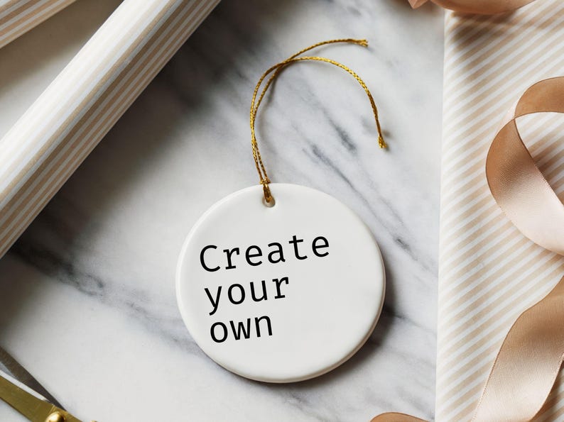 May include: A white, round ceramic ornament with the text "Create your own" in black. The ornament hangs from a gold string. The background includes wrapping paper, ribbon, and a marble surface.