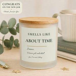 May include: A frosted glass candle with a light wood lid and a label that reads: CONGRATS ON THE NEW JOB Made Just for You, SMELLS LIKE ABOUT TIME, Jenna, Dream job unlocked!, - Your Favorite Sibling. The candle is cylindrical with a pale green label.
