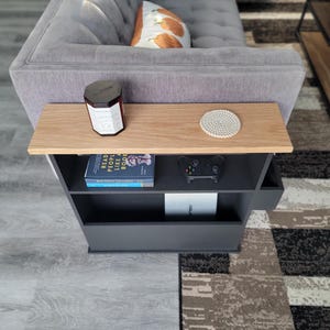May include: A modern side table with a light wood top and a dark gray base. The table has a shelf with a book, a game controller, and a white box. A candle and a coaster sit on the tabletop. The table is next to a gray sofa.