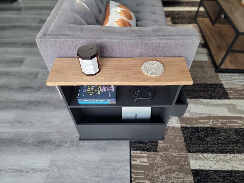 May include: A modern side table with a light wood top and a dark gray base. The table has a shelf with a book, a game controller, and a white box. A candle and a coaster sit on the tabletop. The table is next to a gray sofa.