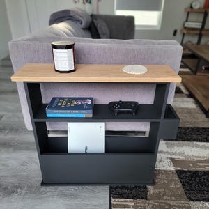May include: A black and wood side table with two shelves and a small storage compartment. The top shelf holds a candle and coaster. The middle shelf has a book and a game controller. The bottom shelf has a white tablet.